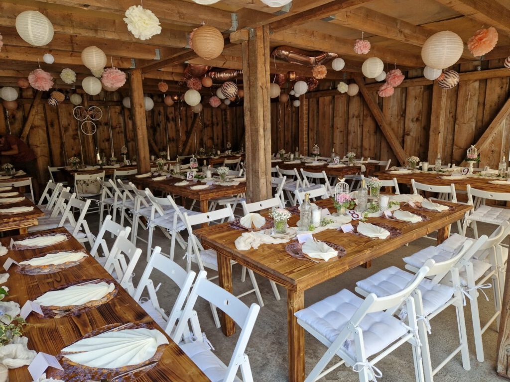 Weiße Klappstühle mit Sitzkissen an Holztischen in einer Scheune mit Lampions